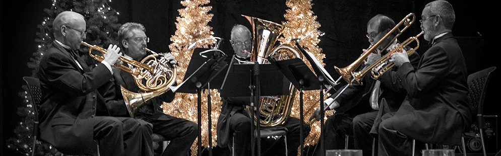 An ensemble of five brass-instrument players sit in front of two gold christmas trees and one other christmas tree on the left. From right to left, the instruments are trumpet, French horn, tuba, trombone, trumpet.