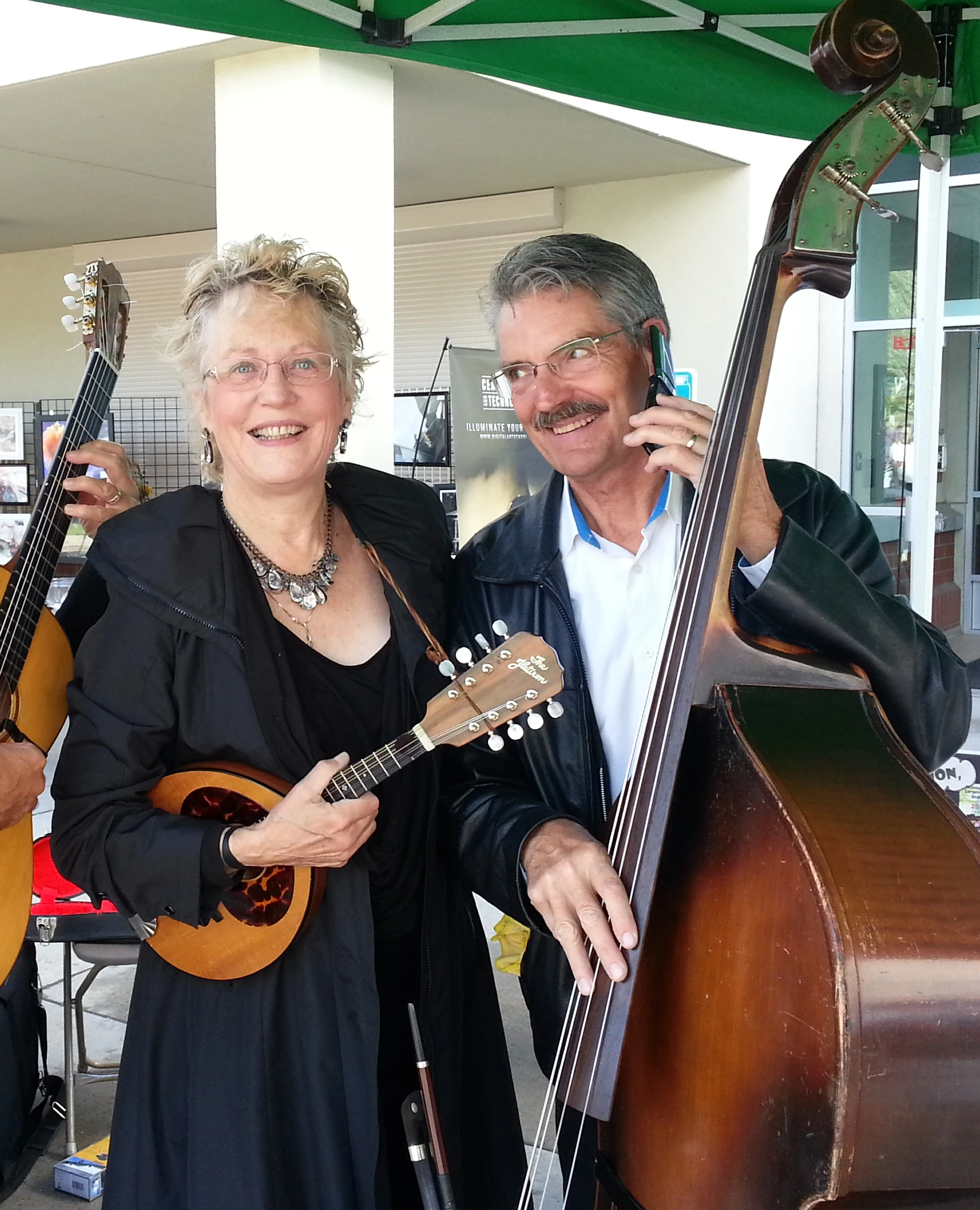 Shari, playing the mandolin, and Edmund, playing the upright bass pose for the camera. Edmund is comedically pretending to talk to someone on a folding cell-phone.