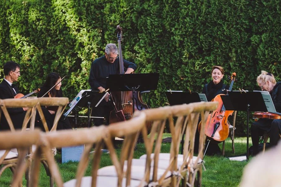 A bass quintet plays in front of a leafy hedge. There are wooden chairs lined up in the foreground.