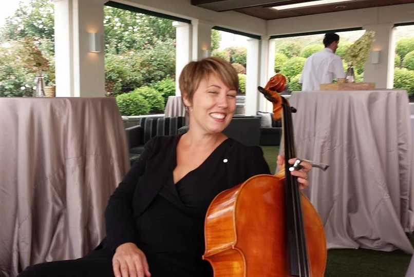 A laughing woman sits with a cello at her side, with two tall, draped tables in the background.