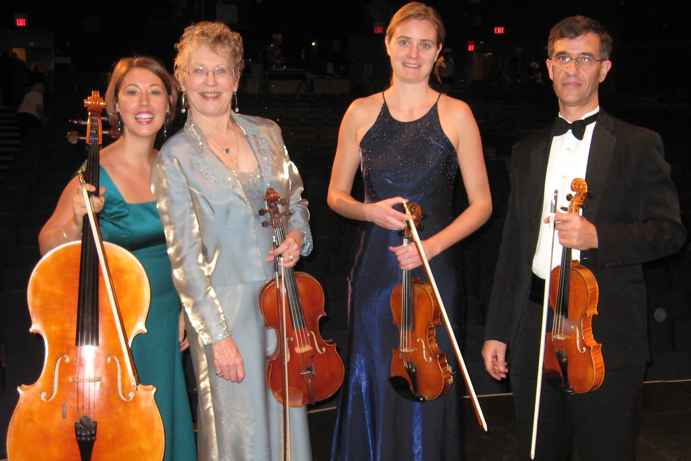 A string quartet in colorful formalwear poses for the camera. They are standing on a stage with a darkened concert hall behind them.