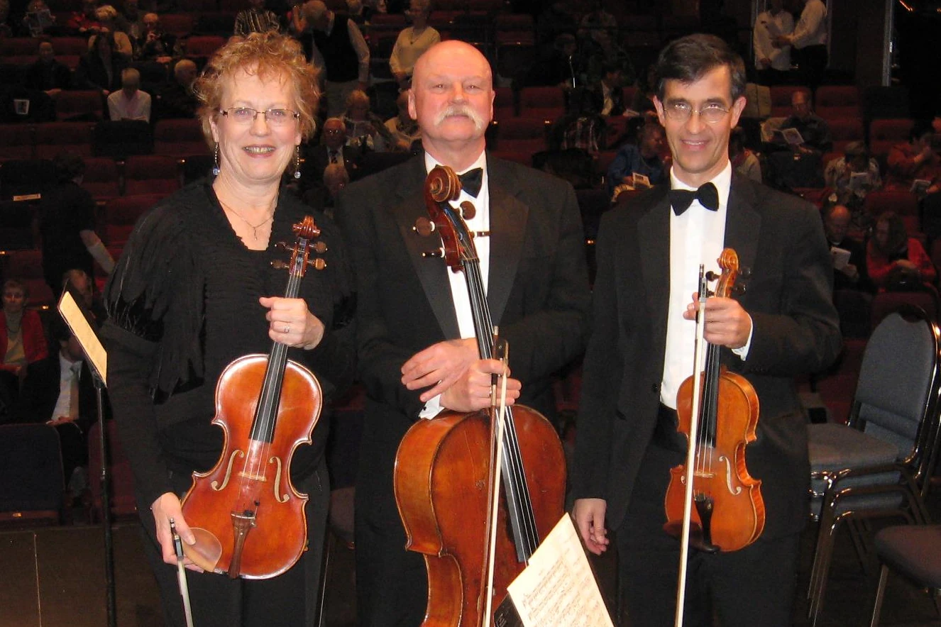 A string trio in dark formalwear and tuxedos for the camera. They are standing on a stage with the audience behind them.