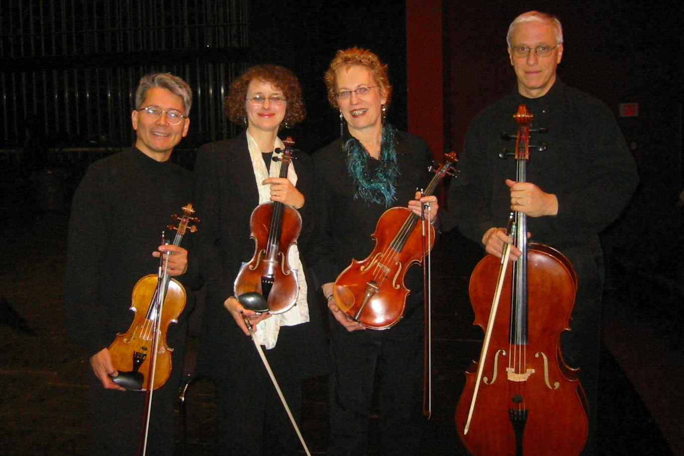 A string quartet in dark formalwear poses for the camera. They are standing on a stage with the audience behind them.