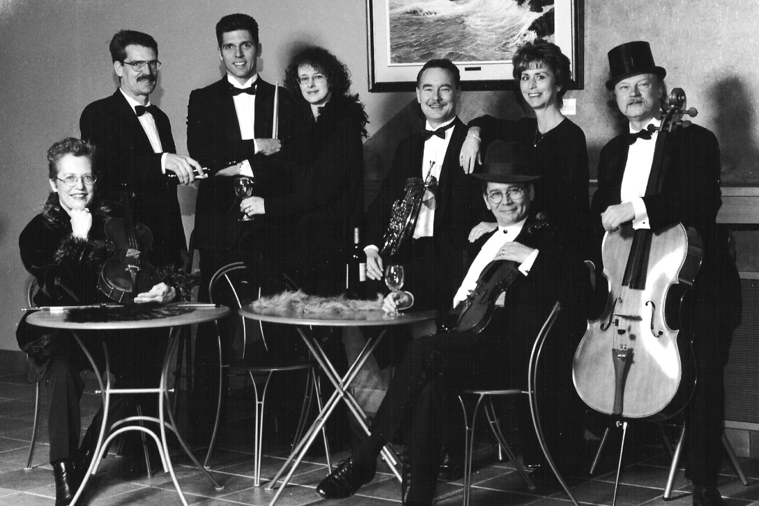 Eight musicians in tuxedos and other formal dress sitting and standing around two tables. One holds a cello.